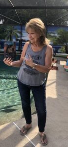 Karen Gehl holds her copy of William Ridley and the Celtic Cross by Connie MacLeod. She is standing poolside, smiling and pointing to the book held in her hand.