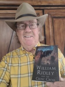 Cousin Ray Ridlon holding his copy of William Ridley and the Celtic Cross by Connie MacLeod. Ray is wearing a yellow and white plaid shirt and wide brimmed cowboy hat, holding the book in front of him.