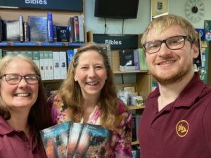 Connie MacLeod - Author with Helen (manager) and Alex at GLO Bookshop in Milngavie. Amid the shelves of books, Helen holds three copies of William Ridley and the Celtic Cross as the trio look forward and smile.