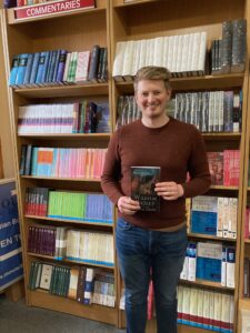 William Frizelle (manager) Evangelical Bookshop - Edinburgh (formerly Mound Books) standing in front of shelves of books holds copy of William Ridley and the Celtic Cross.